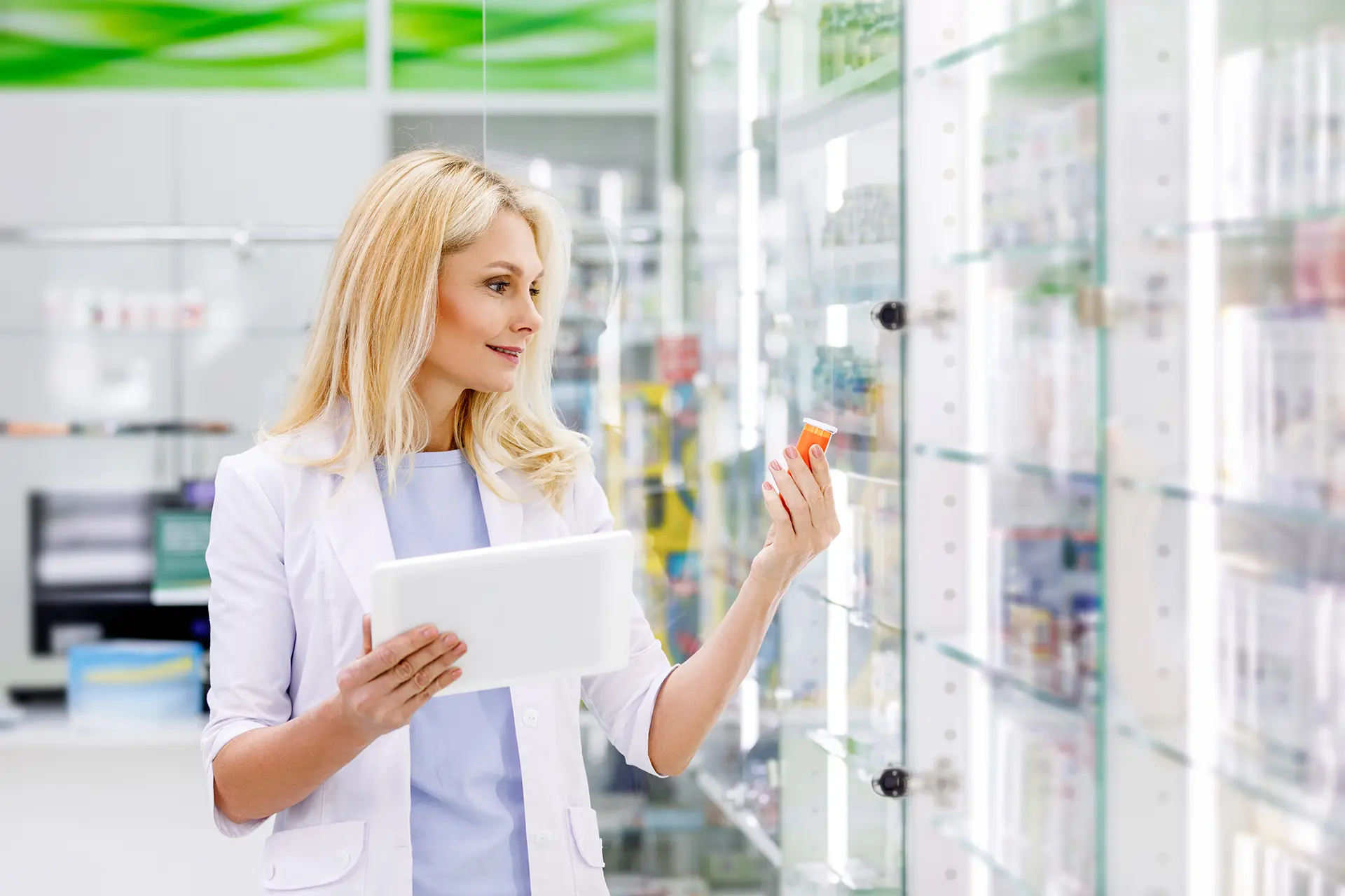 A pharmacist in a white lab coat holds a tablet and examines a medication bottle while standing in front of glass pharmacy shelves stocked with products.
